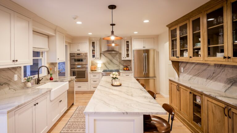 Modern kitchen with white cabinets, marble countertops, a central island, stainless steel appliances, farmhouse sink, wooden chairs, pendant lights, and a rug on the wooden floor.