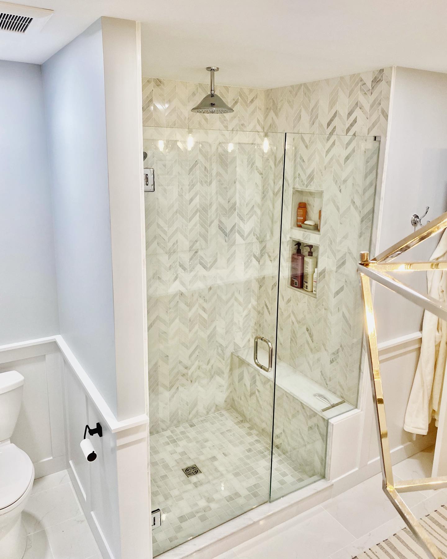 A modern bathroom features a glass-enclosed shower with white herringbone tile walls and a silver showerhead. The room includes a white toilet, a robe on a hook, and a brass towel rack.