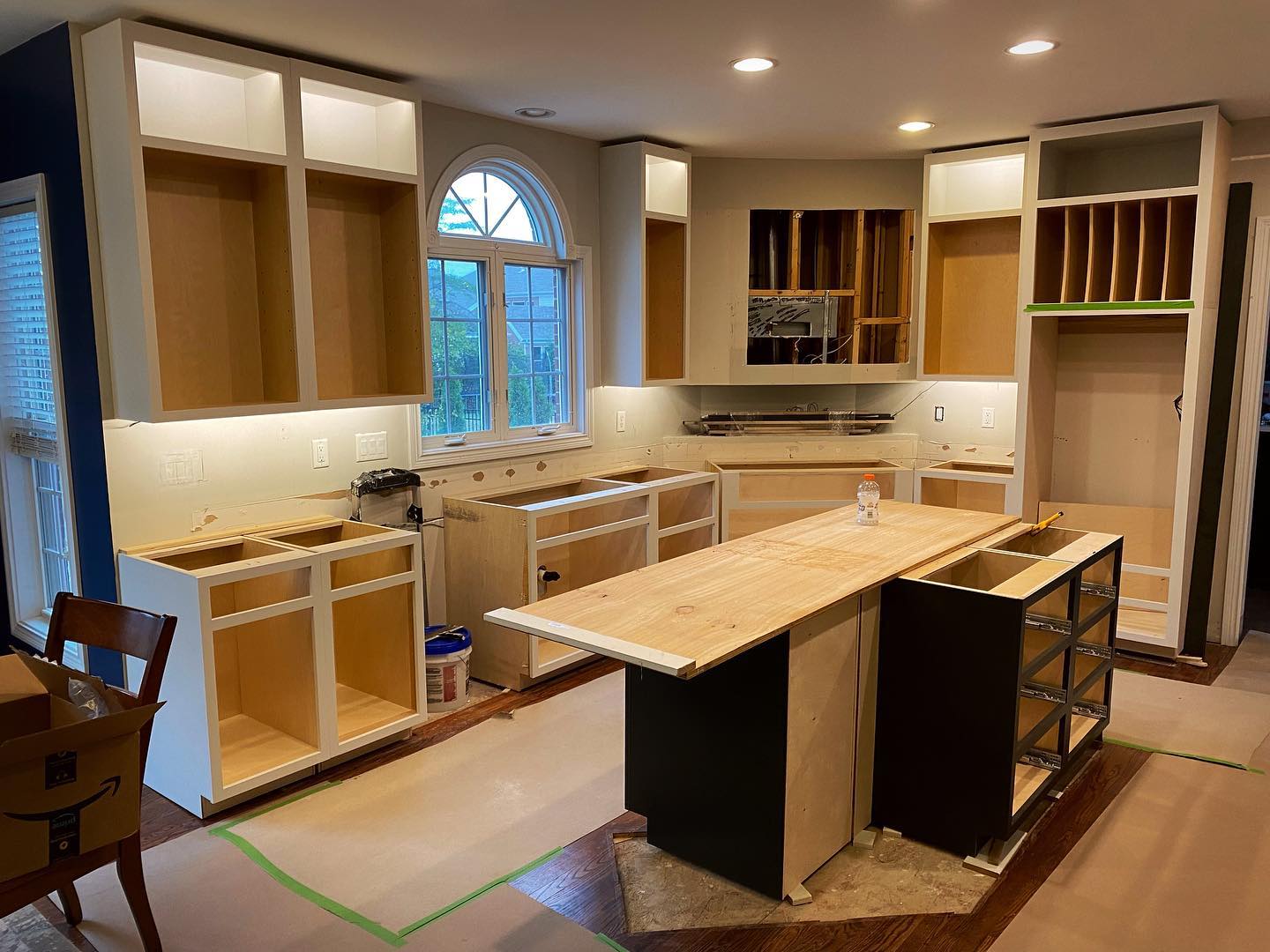 A kitchen under renovation with cabinets partially installed, unfinished center island, and protective coverings on the floor. Some appliances and fixtures are not yet installed.