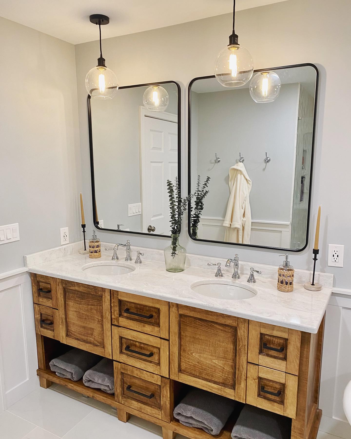 A modern bathroom vanity with a double sink, wooden cabinetry, two mirrors, pendant lights, and a central vase with green foliage. Rolled towels are stored below the sink.