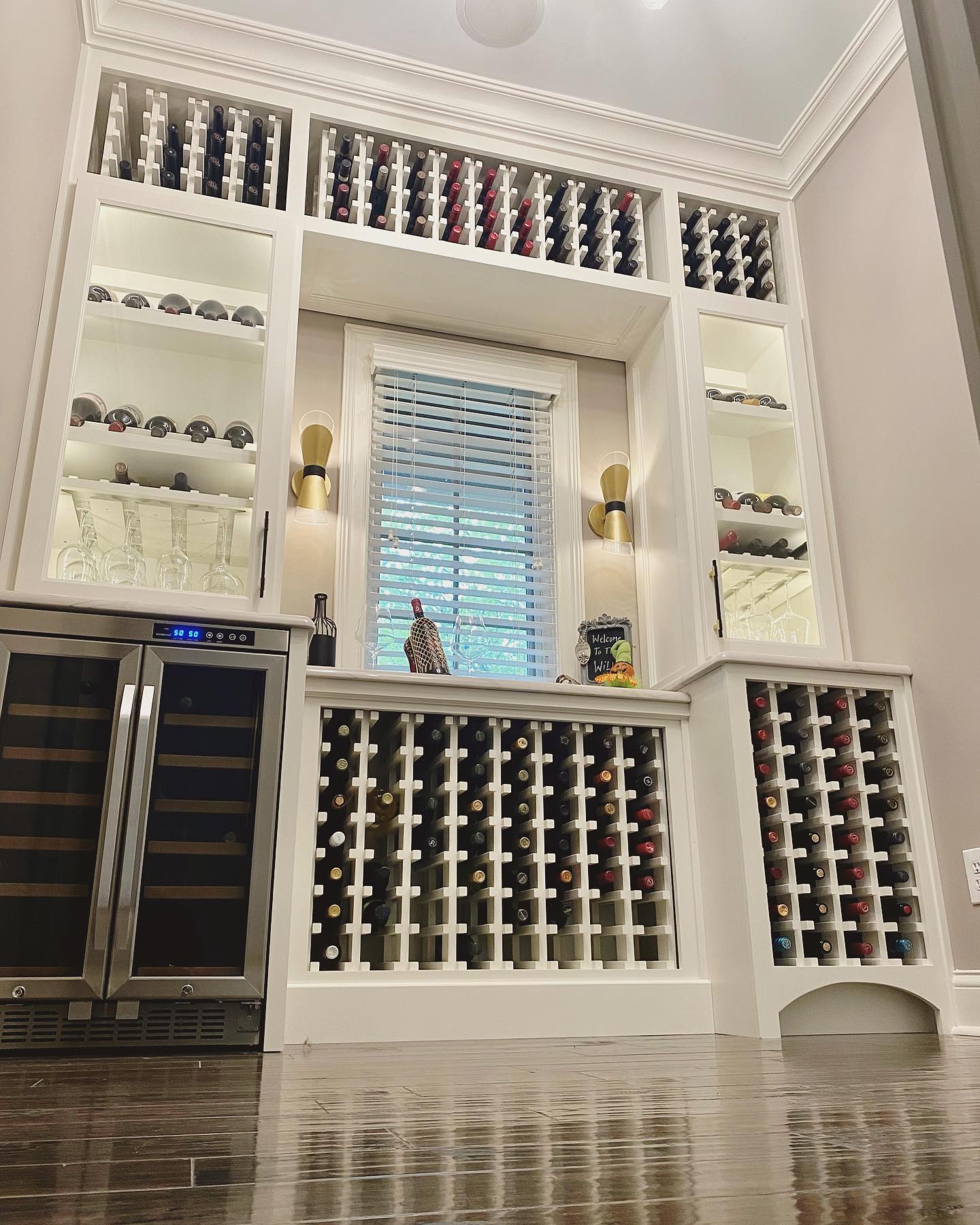 A wine storage room with a window in the center, surrounded by shelves filled with wine bottles, two wall sconces, glass storage, and a wine fridge on the left.