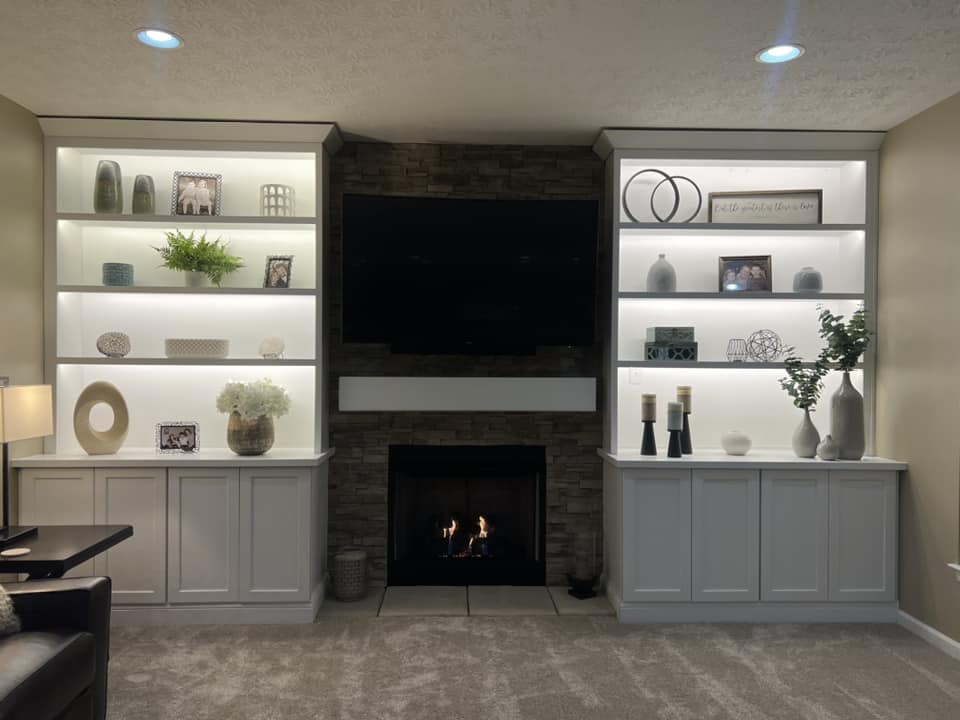 Living room with a built-in fireplace, flanked by two illuminated shelves displaying various decorations, such as vases, framed photos, and plants. A television is mounted above the fireplace.