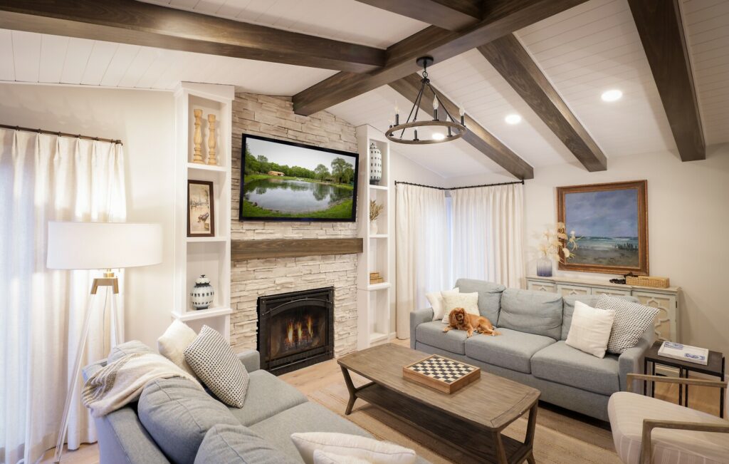 A modern living room featuring a stone fireplace, a TV above it, a chandelier, exposed wooden beams, two gray sofas with cushions, a wooden coffee table with a checkerboard, and a dog resting on a sofa.