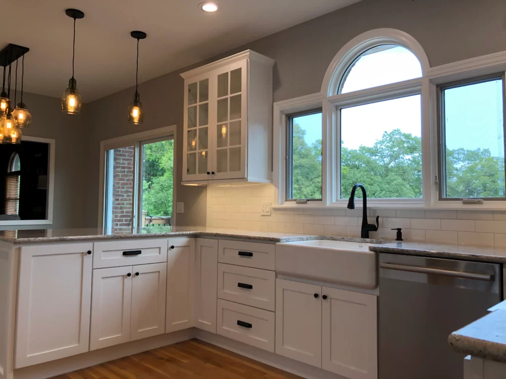 A modern kitchen featuring white cabinets, a farmhouse sink, granite countertops, pendant lights, and large windows with an outdoor view.