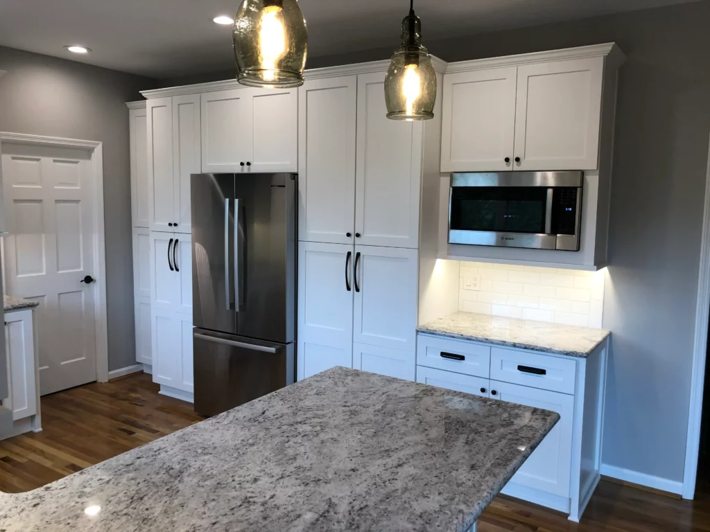 Modern kitchen with white cabinetry, stainless steel refrigerator, microwave, and granite countertops. Two hanging pendant lights illuminate the island counter. Wood flooring is visible.
