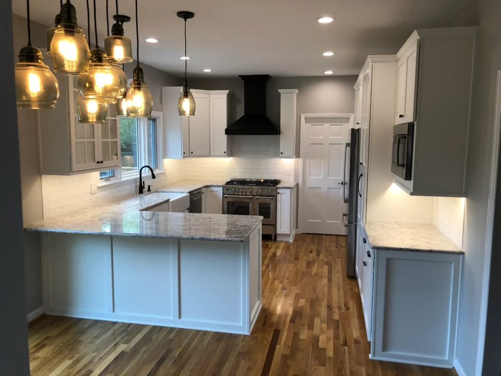Modern kitchen with white cabinets, granite countertops, stainless steel appliances, farmhouse sink, black range hood, and multiple hanging lights over the island. Wooden flooring throughout.
