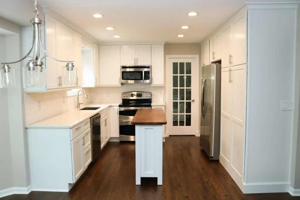 A modern kitchen with white cabinetry, stainless steel appliances, a small central island with a wooden top, dark hardwood flooring, and recessed ceiling lights. A glass-paned door is in the background.