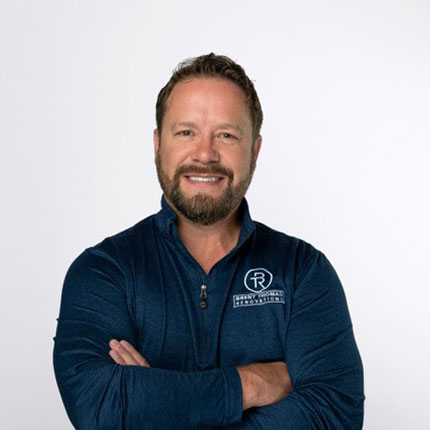 A man with short hair and a beard smiles while crossing his arms, wearing a dark blue pullover with a logo on the left chest. He stands against a plain white background.