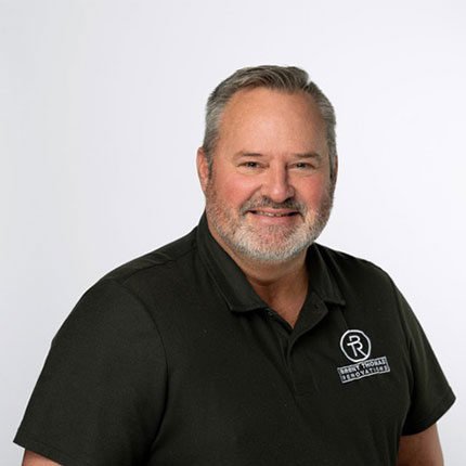 A man with grey hair and a beard wearing a black polo shirt with a company logo, smiling at the camera against a plain white background.