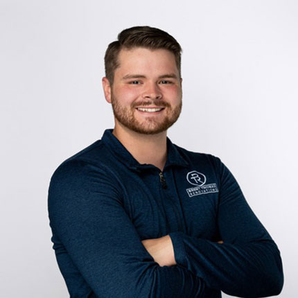 A person with short hair and a beard wearing a dark blue long-sleeve shirt with a logo on it, standing against a plain white background with arms crossed and smiling.