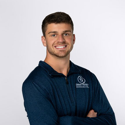 Young man with short dark hair and a beard, wearing a blue zip-up shirt with a logo on his left chest, standing with arms crossed against a white background.