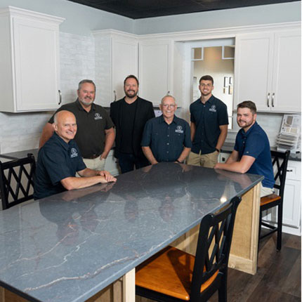 Six men are smiling and posing around a marble-top island in a modern kitchen. White cabinets and light gray walls are visible in the background.